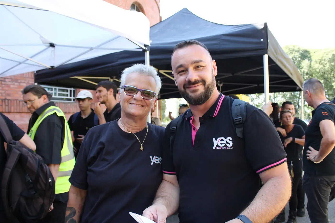 Due collaboratori YES con la maglia e la polo aziendale sorridono sotto il gazebo del punto di ritrovo, prima di un evento